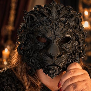May include: A matte black mask shaped like a lion's head, featuring intricate, ornate scrollwork and filigree details throughout the mane and facial features. The mask has two eye openings and covers the upper portion of the face.