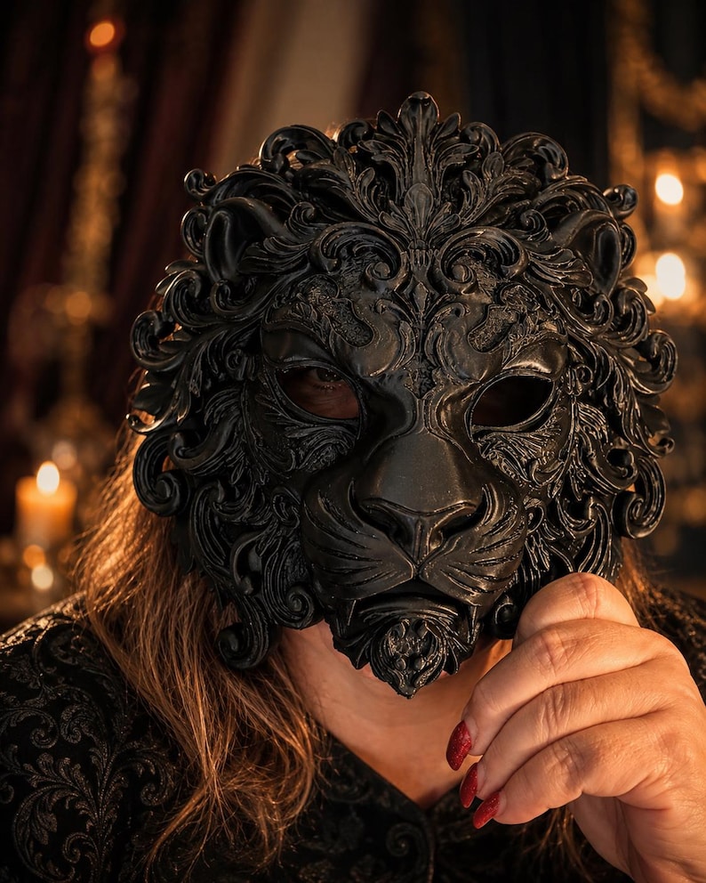 May include: A matte black mask shaped like a lion's head, featuring intricate, ornate scrollwork and filigree details throughout the mane and facial features. The mask has two eye openings and covers the upper portion of the face.
