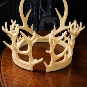 May include: A cream-colored antler crown with multiple points and curved tines. The crown is made of a light-colored material and sits on a dark wooden surface. The crown is a decorative item, possibly for a costume or home decor.