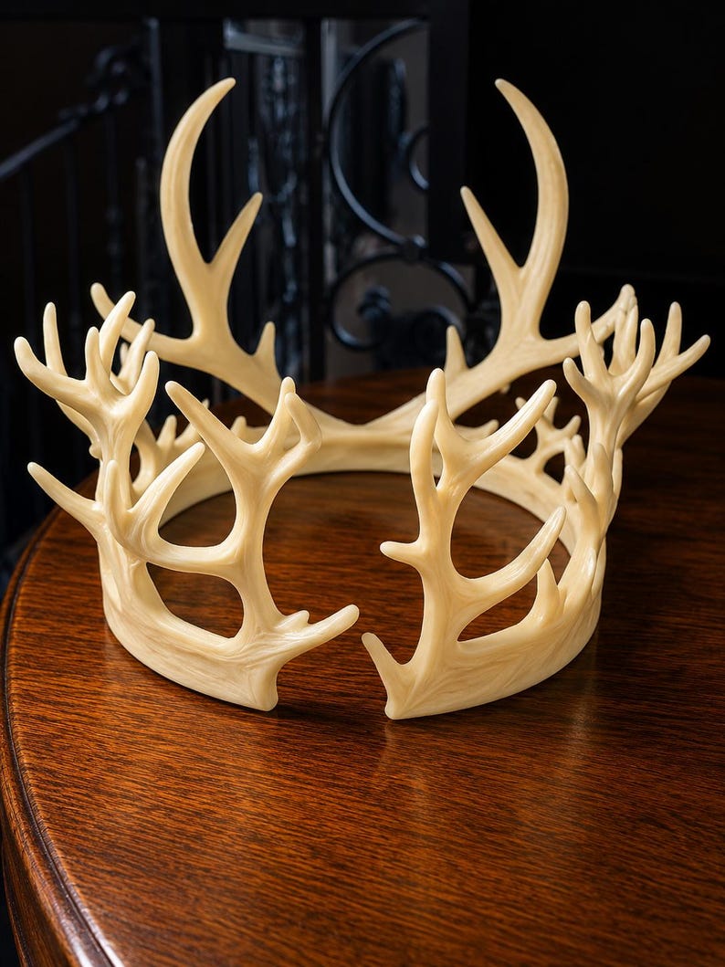 May include: A cream-colored antler crown with multiple points and curved tines. The crown is made of a light-colored material and sits on a dark wooden surface. The crown is a decorative item, possibly for a costume or home decor.