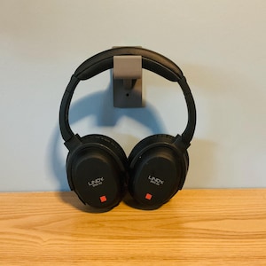 May include: Black over-ear headphones hanging on a gray wall mount. The headphones have the brand name "LINDY" printed on the earcups, with a small red square. The headphones are resting on a wooden surface.