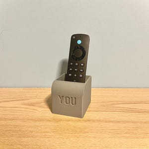 May include: A gray, cube-shaped remote control holder with the word "YOU" embossed on the front. A black remote control is inside the holder. The holder is on a wooden surface.