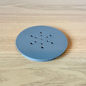 Może przedstawiać: Okrągły, jasnoniebieski przedmiot z wieloma małymi otworami ułożonymi we wzór. Obiekt wykonany jest z litego materiału i leży na jasnobrązowej drewnianej powierzchni. Średnica: około 10 cm.