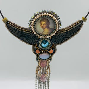 May include: Handmade beaded necklace featuring a central portrait of a woman, surrounded by pearls and crystals. The necklace has dark green wings, a large blue crystal, and dangling chains with pink and clear stones. The necklace is on a black cord.
