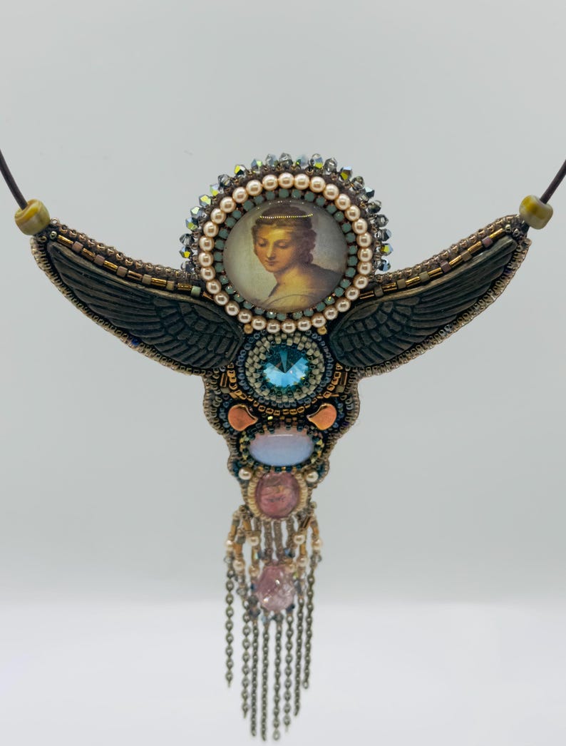 May include: Handmade beaded necklace featuring a central portrait of a woman, surrounded by pearls and crystals. The necklace has dark green wings, a large blue crystal, and dangling chains with pink and clear stones. The necklace is on a black cord.