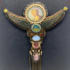 May include: A handcrafted necklace with a central portrait, surrounded by a halo of beads. The necklace features bronze-colored wings, various gemstones, and a cascading fringe. The design incorporates shades of gold, teal, pink, and cream.