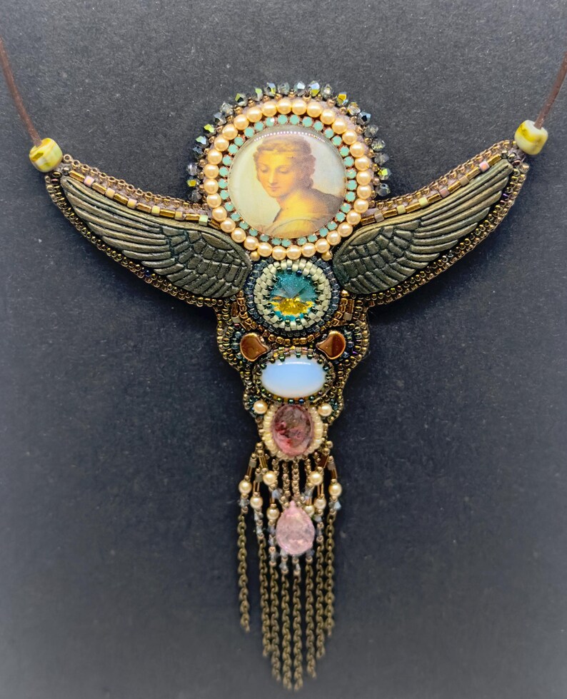 May include: A handcrafted necklace with a central portrait, surrounded by a halo of beads. The necklace features bronze-colored wings, various gemstones, and a cascading fringe. The design incorporates shades of gold, teal, pink, and cream.