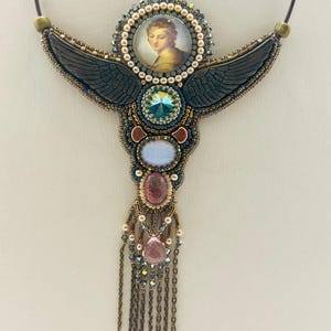 May include: A handcrafted necklace featuring a central portrait surrounded by a beaded halo. Dark gray wings extend from the center, adorned with various gemstones in blue, pink, and clear hues. The necklace has a chain fringe.