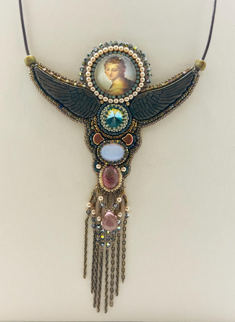 May include: A handcrafted necklace featuring a central portrait surrounded by a beaded halo. Dark gray wings extend from the center, adorned with various gemstones in blue, pink, and clear hues. The necklace has a chain fringe.