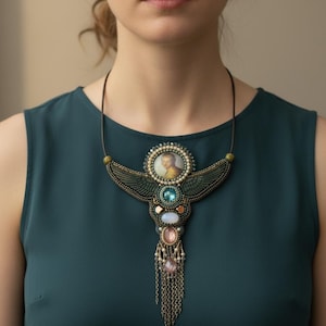 May include: A handcrafted statement necklace featuring a central portrait within a beaded frame. The pendant showcases a winged design with various gemstones in blue, pink, and white, accented with dangling chains. The necklace is worn on a dark green top.