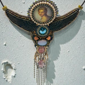 May include: Ornate, handcrafted necklace featuring a central portrait of a woman, surrounded by beads and gemstones. The design incorporates dark wings, a large blue gem, and dangling chains with pink and clear stones. The necklace is suspended from a dark cord.