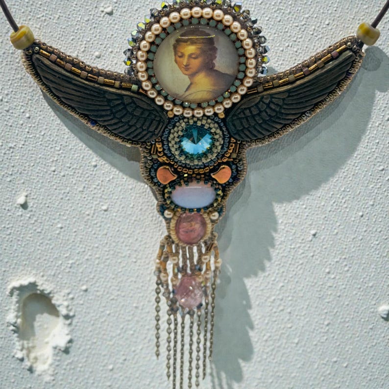 May include: Ornate, handcrafted necklace featuring a central portrait of a woman, surrounded by beads and gemstones. The design incorporates dark wings, a large blue gem, and dangling chains with pink and clear stones. The necklace is suspended from a dark cord.