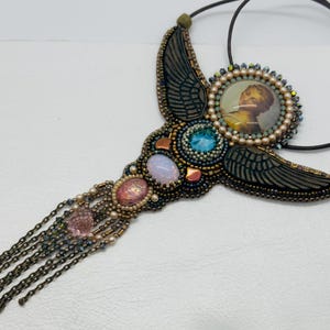 May include: A handcrafted necklace featuring a central image of a person, surrounded by beads and gemstones. The necklace has a wing-like design with a mix of blue, pink, and gold accents, and a chain fringe. The necklace is on a black cord.