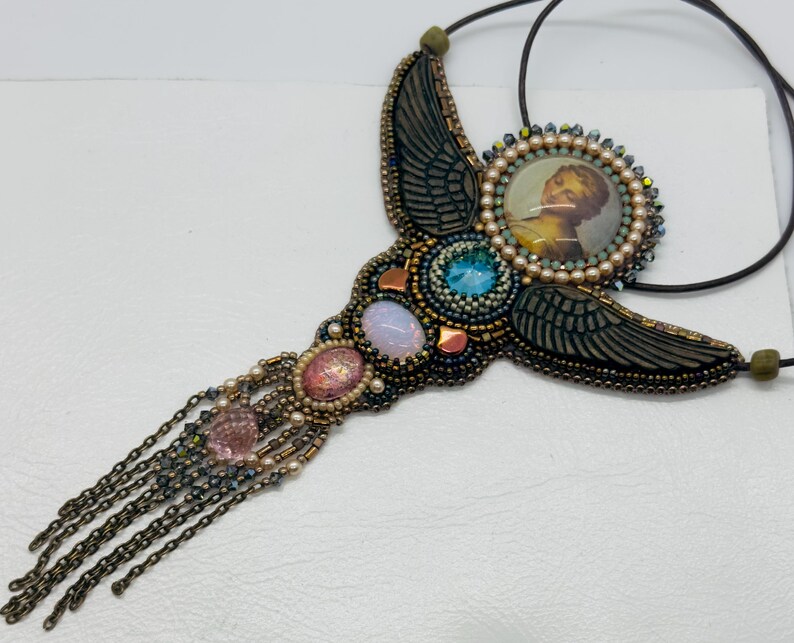 May include: A handcrafted necklace featuring a central image of a person, surrounded by beads and gemstones. The necklace has a wing-like design with a mix of blue, pink, and gold accents, and a chain fringe. The necklace is on a black cord.