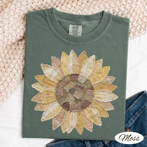 May include: A moss green t-shirt featuring a large sunflower applique. The sunflower is made of various fabric patches in shades of yellow, tan, and brown. The shirt has a crew neck and short sleeves. The brand tag "Comfort Colors" is visible at the neckline.