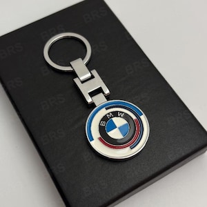 May include: A silver-toned metal keychain featuring a circular BMW logo emblem with blue, white, and red enamel accents. The keychain has a standard split ring and a rectangular connector piece, presented on a black rectangular box with BRS text.