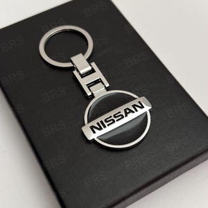 Pode incluir: Um porta-chaves de metal prateado com um emblema circular que apresenta a palavra NISSAN gravada em letras pretas no centro. O porta-chaves inclui um anel padrão fixado a uma peça de conexão retangular, tudo com um acabamento polido e brilhante.