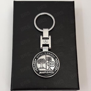 Könnte beinhalten: Ein silberfarbener Metallschlüsselanhänger mit einem kreisförmigen Emblem, das den Text AFFALTERBACH und AMG trägt. Das Emblem zeigt einen Baum und eine Ventilgrafik. Ein kleines Mercedes-Benz-Logo ist auf dem rechteckigen Verbindungsstück eingraviert.