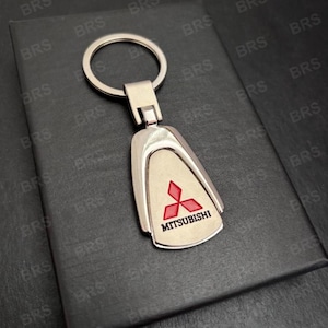 Könnte beinhalten: Ein silberfarbener Metall-Schlüsselanhänger mit einem tropfenförmigen Anhänger. Der Anhänger zeigt das rote Mitsubishi-Logo und den Markennamen in Schwarz. Der Schlüsselanhänger hat einen silbernen Ring zum Befestigen von Schlüsseln.