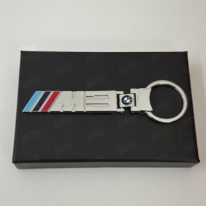 Può includere: Un portachiavi in metallo color argento con il logo M3 e strisce diagonali blu chiaro, blu scuro e rosse sul lato sinistro. Un piccolo emblema circolare BMW è posizionato tra la scritta M3 e l'anello portachiavi circolare.