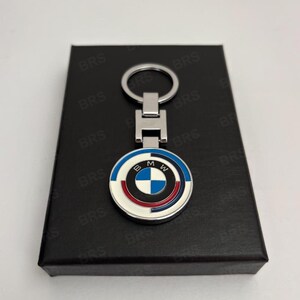 May include: A silver-toned metal keychain featuring a circular pendant with the BMW logo in the center, surrounded by concentric rings in blue, white, and red. The pendant is attached to a rectangular metal link and a circular split ring.