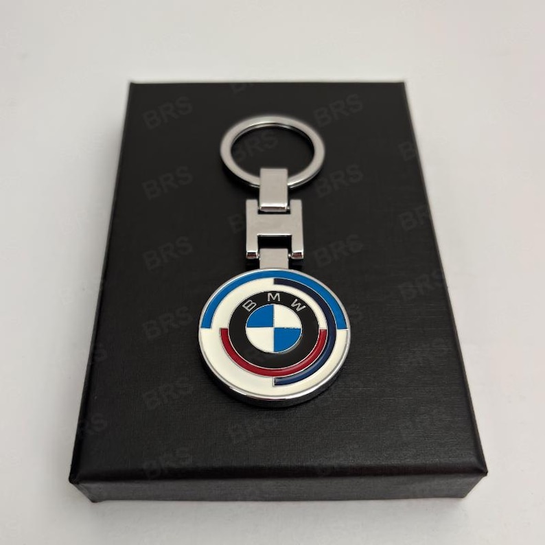 May include: A silver-toned metal keychain featuring a circular pendant with the BMW logo in the center, surrounded by concentric rings in blue, white, and red. The pendant is attached to a rectangular metal link and a circular split ring.