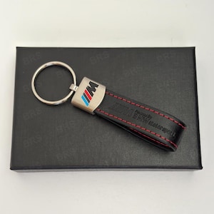 Może przedstawiać: Czarny skórzany brelok do kluczy ze srebrnym metalowym mocowaniem z logo BMW M w kolorze niebieskim, fioletowym i czerwonym. Skórzany pasek ma czerwone przeszycia i wytłoczony napis BMW Motorsport. Brelok posiada okrągłe metalowe kółko.