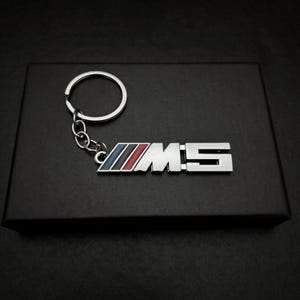 Könnte beinhalten: Ein silberner Metall-Schlüsselanhänger mit den Buchstaben "M5". Der Schlüsselanhänger hat einen Ring und eine Kette. Das "M5"-Logo hat blaue, rote und schwarze Streifen. Der Schlüsselanhänger liegt auf einer schwarzen Box.