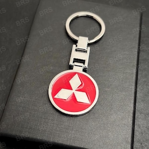 Pode incluir: Um porta-chaves prateado com um emblema circular vermelho com um logótipo branco de três diamantes. O porta-chaves tem um anel e um conector retangular. O fundo é uma superfície escura e texturizada.