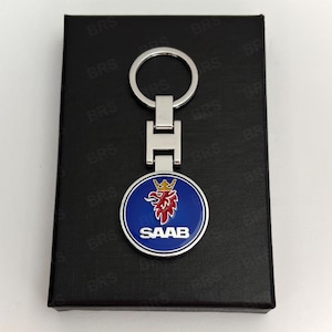 Peut inclure: Un porte-clés en métal argenté doté d'un emblème circulaire avec un fond bleu, une tête de griffon rouge avec une couronne dorée et le texte blanc SAAB. Le porte-clés présente une finition polie et inclut un anneau fendu rond standard.