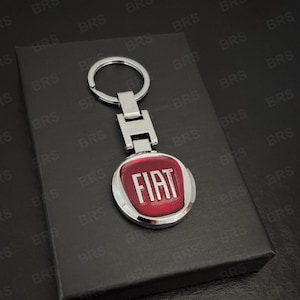 May include: A silver-toned metal keyring featuring a circular pendant with a red background and the word FIAT in raised silver lettering. The keyring includes a standard split ring attached to a rectangular metal connector piece above the logo.