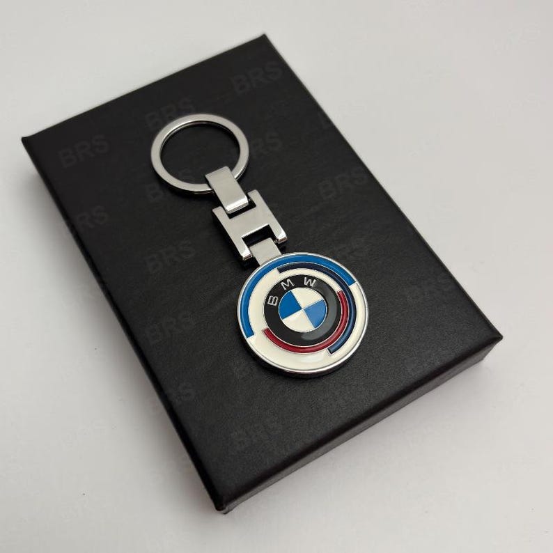 May include: A silver-toned metal keychain featuring a circular BMW logo emblem with blue, white, and red enamel accents. The keychain has a standard split ring and a rectangular connector piece, presented on a black rectangular box with BRS text.