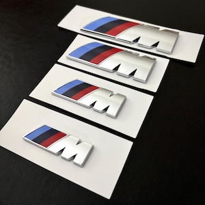 Könnte beinhalten: Vier verchromte "M" Embleme mit blau, schwarz und rot gestreiften Streifen, jeweils auf einem weißen rechteckigen Träger. Die Embleme sind diagonal auf einer dunklen Oberfläche angeordnet und weisen eine glänzende Oberfläche und ein modernes Design auf.
