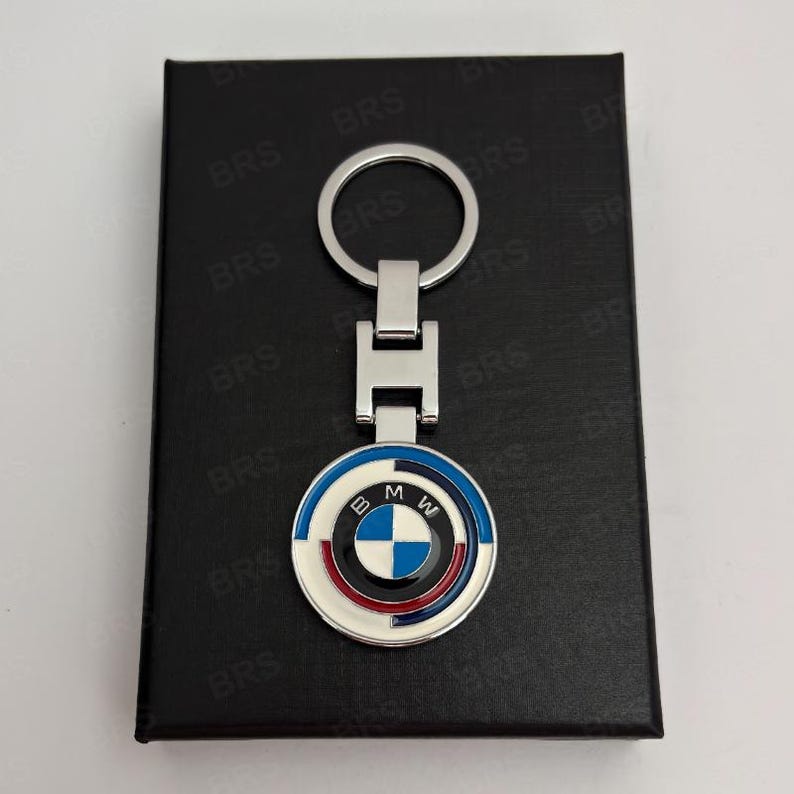 May include: A silver-toned metal keychain featuring a circular BMW logo emblem with blue, white, and red enamel detailing. The circular pendant is attached to a rectangular metal link and a standard round split ring for keys.