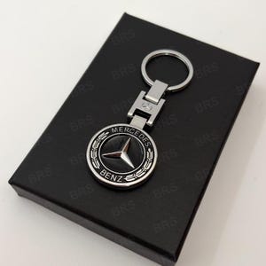 May include: A silver-toned metal keyring featuring a circular emblem with the Mercedes-Benz logo in the centre, surrounded by a laurel wreath design and the text MERCEDES BENZ. The keyring includes a split ring and a rectangular connector piece.