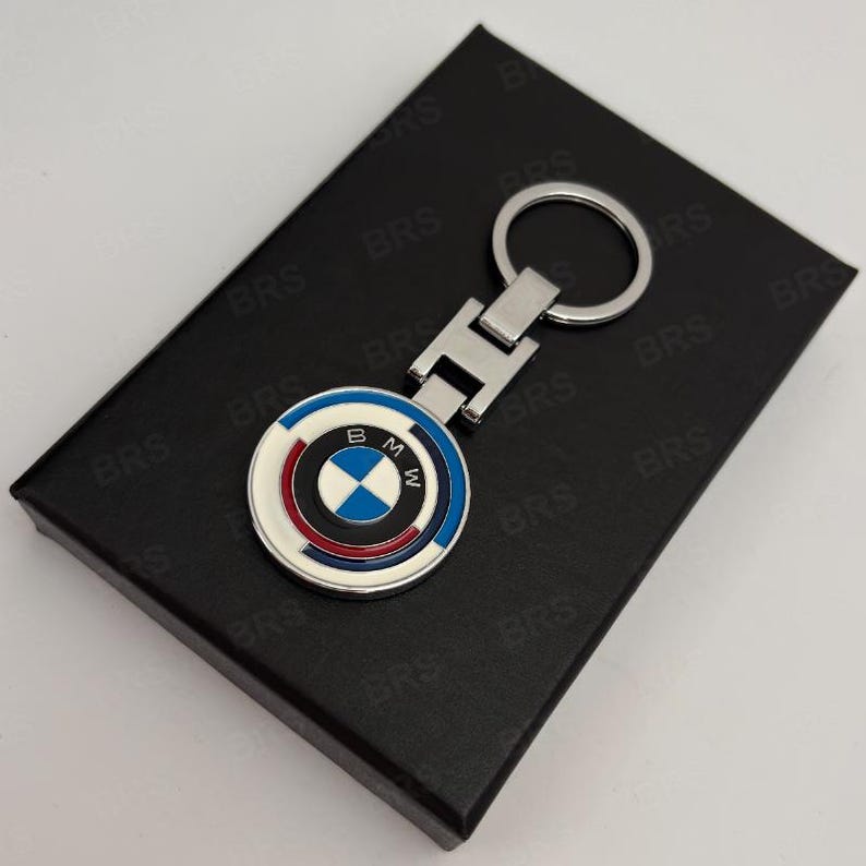 May include: A silver-toned metal keychain featuring a circular emblem with the BMW logo in the center, surrounded by concentric rings of white, blue, and red enamel. The keychain includes a rectangular connector piece and a standard round ring.