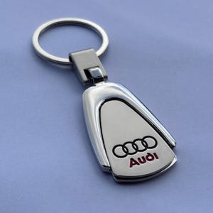 Peut inclure: Un porte-clés Audi argenté avec un anneau. Le porte-clés a une forme rectangulaire avec un centre blanc et le logo Audi en noir avec du texte rouge. Le porte-clés est en métal et a une finition brillante.