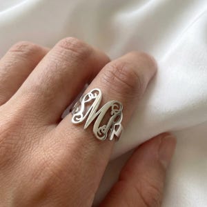 Puede incluir: Un anillo de monograma plateado con las letras "M" y "R" en un diseño calado. El anillo se lleva en un dedo, mostrando su estética elegante y personalizada. El anillo está hecho de un metal brillante.