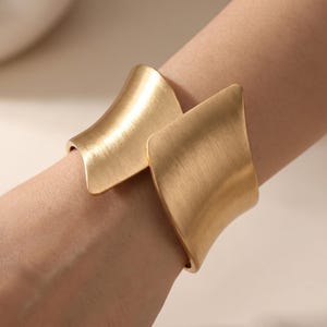 Könnte beinhalten: Ein goldfarbenes Armreif-Armband mit gebürsteter Metalloberfläche. Das Armband hat ein einzigartiges Design mit zwei gebogenen, rechteckigen Paneelen, die sich leicht überlappen. Das Armband wird am Handgelenk getragen.