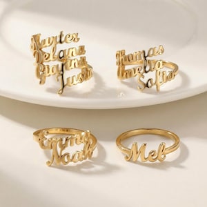 Puede incluir: Cuatro anillos dorados con nombres, presentados en un plato blanco. Los anillos presentan nombres en escritura cursiva: "Hunter, Delana, Grayson, Justin", "Thomas, Amelia, Sofia", "Emma, Noah" y "Mel".