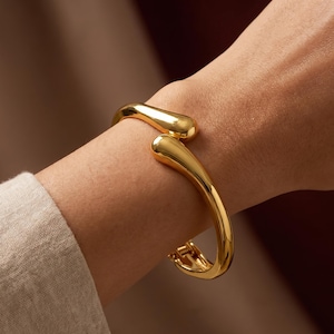 Puede incluir: Una pulsera de puño dorada con un diseño curvo y abierto. La pulsera tiene un acabado liso y pulido y un cierre de broche. El diseño presenta extremos redondeados en forma de lágrima. La joya se muestra en la muñeca de una persona.