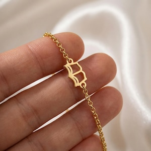 Puede incluir: Una pulsera de cadena dorada con un colgante con la letra "B". La pulsera es delicada, con una cadena fina y un pequeño colgante calado. El fondo es de un color suave y neutro, resaltando los detalles de la joya.