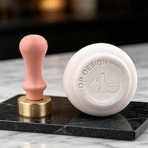 Puede incluir: Un sello de lacre rosa con base de latón y un sello de cerámica blanco con el texto "DP DESIGN" y un diseño de jarrón y cuenco. Los artículos están sobre una superficie de mármol negro.