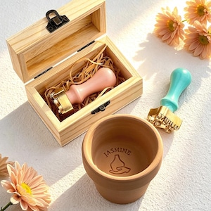 May include: A wooden box containing a pink stamp with a gold stamp head, a turquoise stamp, and a terracotta pot with the word "Jasmine" and a graphic. The box is open. The pot is empty.