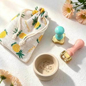May include: A selection of craft items: a drawstring bag with a pineapple pattern, a small ceramic bowl with a cat design and text, and two stamps with wooden handles and gold-coloured stamp heads. The stamps have teal and pink colours.