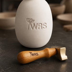 May include: A white oval ceramic object featuring an embossed dandelion graphic and the letters WRS. Beside it lies a wooden handle stamp with a brass head, also displaying the same dandelion graphic and the letters WRS engraved on the wood.