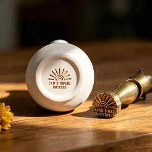 Op de afbeelding: Een witte keramische aardewerken basis met een gestempeld zonlogo en de tekst JAMIE DUVAL POTTERY, geplaatst naast een messing stempelgereedschap dat hetzelfde zonlogo en omgekeerde tekst toont voor branding.