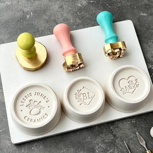Könnte beinhalten: Eine Sammlung von Keramikstempeln und passenden Schalen. Die Stempel haben farbige Griffe in Gelb, Rosa und Türkis, mit goldfarbenen Metallstempelköpfen. Die weißen Schalen sind mit eingravierten Designs und Texten versehen, darunter die Worte "Lessie June's Ceramics."