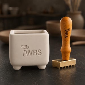 May include: A square, off-white ceramic planter with four small feet and the text WRS with a dandelion graphic embossed on the front, positioned next to a wooden-handled metal stamp featuring the same WRS text and dandelion graphic.