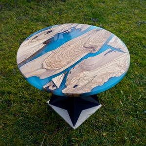 Può includere: Un tavolino rotondo dal design unico. Il piano del tavolo presenta una combinazione di legno chiaro e resina blu brillante. Il tavolo è sostenuto da una base geometrica bianca e nera, creando un'estetica moderna. Il diametro è di circa 76 cm.